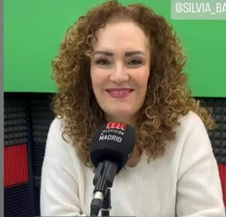 Mujer con cabello rizado y sonrisa, sentada frente a un micrófono en estudio de radio.
