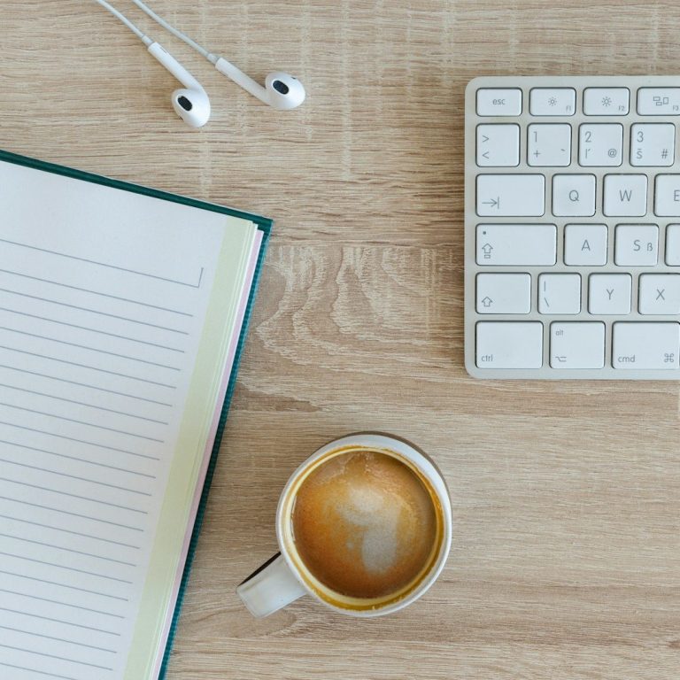 Taza de café, cuaderno, teclado y auriculares sobre una mesa de madera.