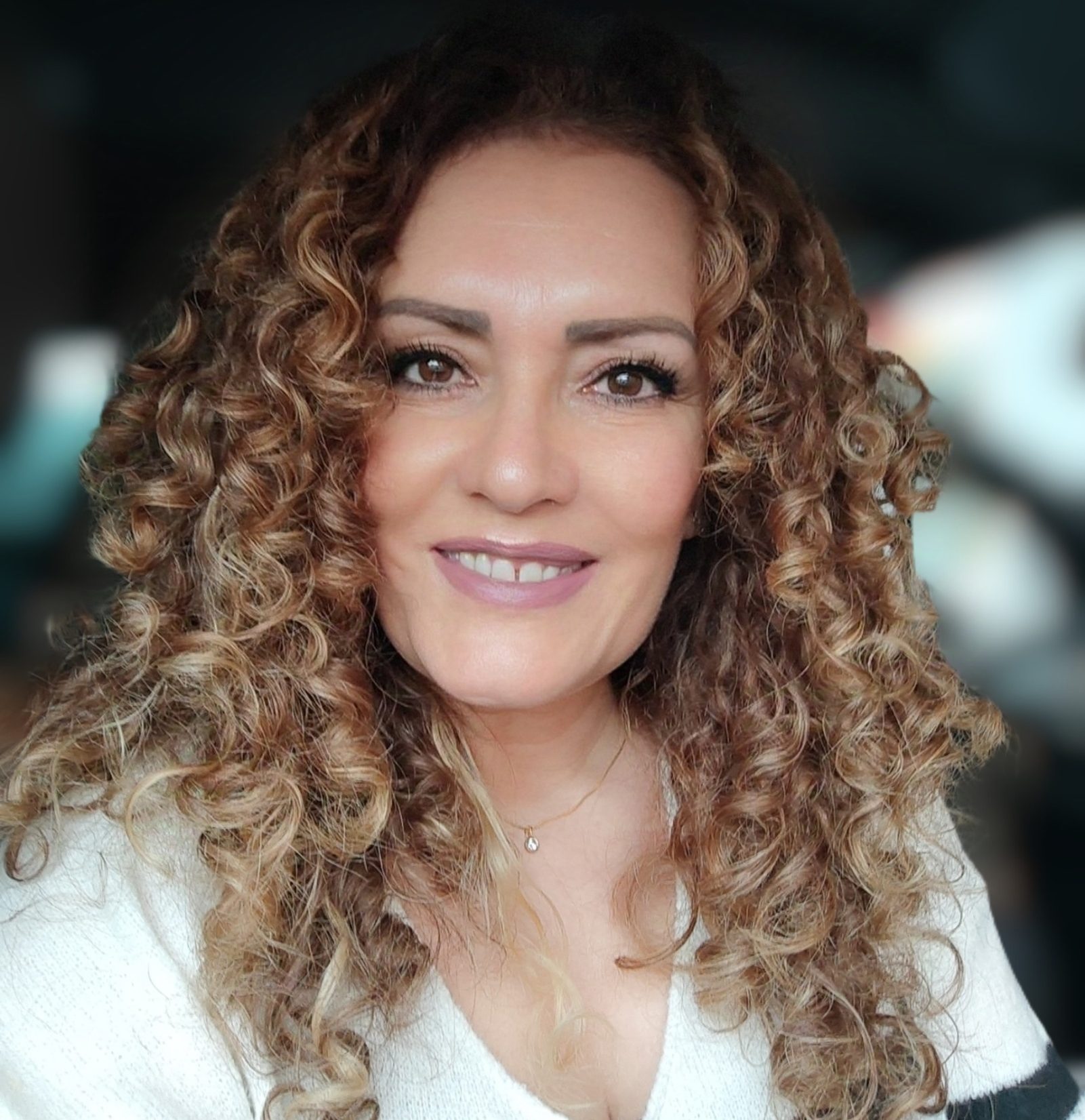 Mujer con cabello rizado y rubio, sonriente, en un entorno con luz tenue.