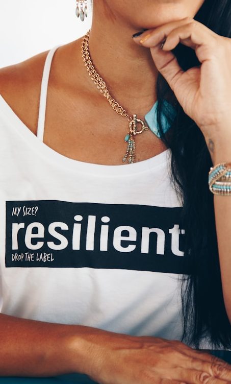 Camisa blanca con la palabra "resilient" en negro, joyas y un fondo desenfocado.